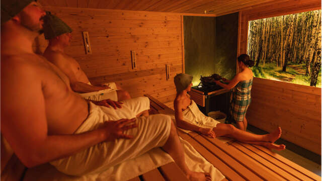 Gäste in der Birkensauna, Aufgießerin mit Birkenzweigen vor beleuchtetem Bild mit Birkenwald